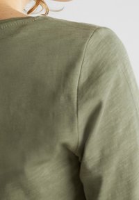 Oliwkowo-zielony, lekki, teksturowany materiał z okrągłym dekoltem i widocznym przeszyciem. Posiada luźny krój i gładką powierzchnię.