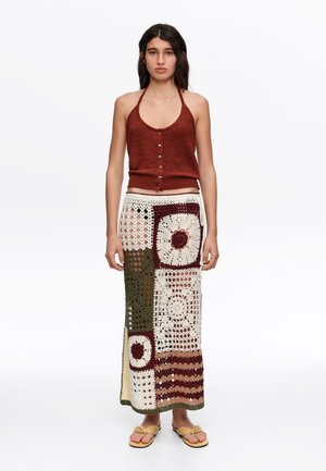 Mujer con top de punto sin mangas de color óxido con botones y una falda larga de crochet patchwork en tonos crema, marrón y verde, de pie descalza.