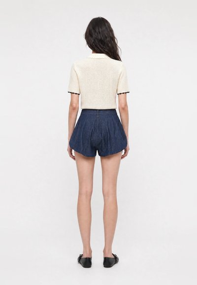 Femme de dos portant un top en maille blanche à manches courtes avec bordure noire et un short plissé bleu foncé taille haute avec des chaussures noires.