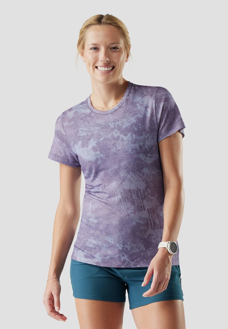 Mujer sonriendo con una camisa de manga corta de color púrpura con estampado, pantalones cortos azul verdoso y un smartwatch blanco en su muñeca izquierda, sobre un fondo liso.