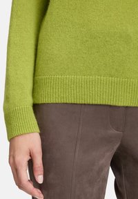 Ręka opierająca się na boku, ubrana w limonkowy sweter z dzianiny i brązowe zamszowe spodnie z widocznym szyciem.