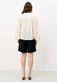 Spitzenbluse mit langen Ballonärmeln in Creme, kombiniert mit schwarzen Shorts. Die Bluse zeichnet sich durch ein florales Muster und ein lockeres, fließendes Design aus.