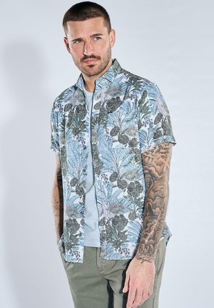 Mann mit kurzen dunklen Haaren, der ein blau-graues kurzärmeliges Hemd mit Blumenmuster über einem weißen T-Shirt und grünen Hosen trägt, mit tätowierten Armen, schaut nach links.