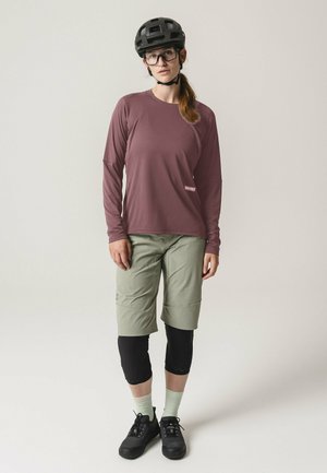 Frau trägt schwarzen Fahrradhelm, Brille, mauvefarbenes Langarmshirt, hellgrüne Shorts über schwarzen Leggings und schwarze Schuhe, steht vor einfarbigem Hintergrund.