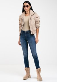 Beige prošívaná bunda s límcem, žebrovanými manžetami a předními kapsami; nošena přes přiléhavý béžový top a tmavě modré skinny džíny; béžové boty.