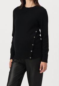 Woman wearing a black long-sleeve sweater with side buttons and black leather pants, standing against a plain light background.