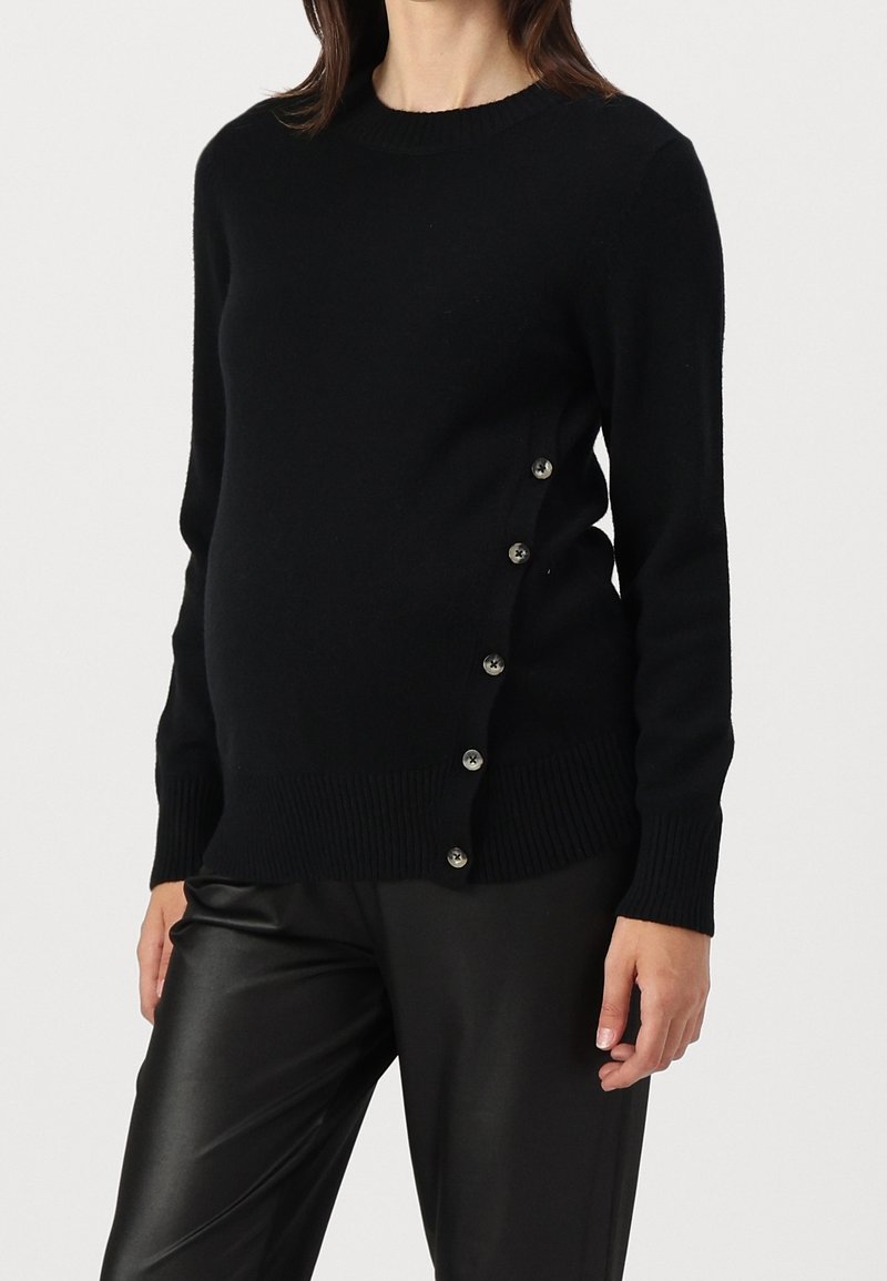 Woman wearing a black long-sleeve sweater with side buttons and black leather pants, standing against a plain light background.