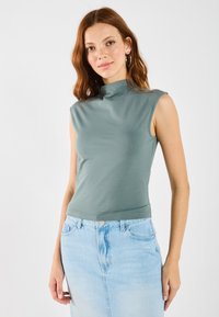 Tett, ermeløs høyhalset topp i dempet grønn stoff, kombinert med lys blå denimskjørt med frontsplitt og klassiske knappedetaljer.