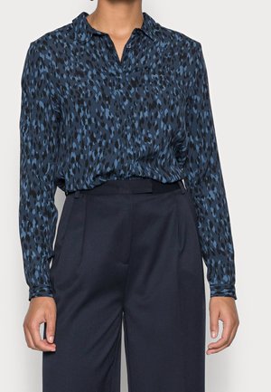 Femme portant un chemisier bleu et noir à manches longues à motifs, rentré dans un pantalon taille haute bleu marine foncé, sur un fond uni.