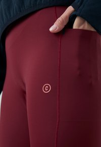 Primer plano de una mano tirando de la cinturilla de unas mallas deportivas de color rojo oscuro con un pequeño logo de círculo "C" en el muslo.