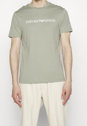 Homme portant un t-shirt vert clair Emporio Armani et un pantalon crème, debout avec les bras détendus le long du corps sur fond blanc.