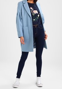 Ljusblå, oversized kappa med en enda ficka, bärs över en marinblå skjorta med blommorbroderi, kombinerad med mörka jeans och vita sneakers.