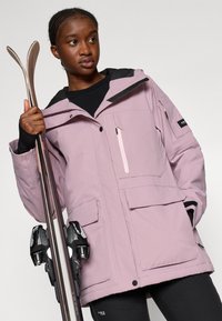 Világos rózsaszín síkabát látható cipzárakkal és zsebekkel, textúrált fekete kapucnival, fekete sínadrággal viselve. Sífelszerelés az egyik oldalon.