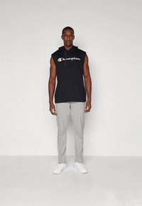 Mann som står, iført svart ermeløs Champion-hettegenser, lysegrå joggebukser og hvite joggesko mot en ensfarget lys bakgrunn.