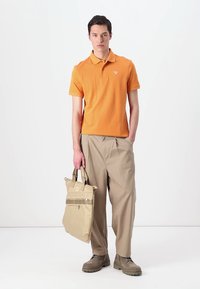 Πορτοκαλί πόλο μπλούζα, μπεζ καρό παντελόνια και καφέ υπόδηση. Κρατώντας μια μπεζ τσάντα tote με φερμουάρ και διακοσμητικά στοιχεία. Απλή, casual εμφάνιση.