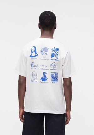 Mand iført hvid t-shirt med blå kunstneriske portrætter og navne på berømte malere på ryggen, stående mod en ensfarvet baggrund.