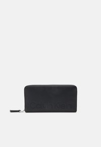 Crna kožna novčanica sa zatvaračem na patentni zatvarač. Sadrži perforirani logo "Calvin Klein" sprijeda i glatku teksturu. Tanak, pravokutni oblik.