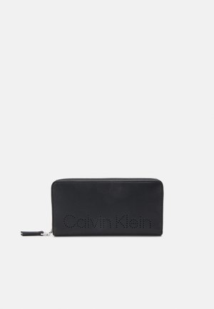 Black leather wallet with a zip closure. Features perforated "Calvin Klein" logo on the front and a smooth texture. Slim, rectangular shape.