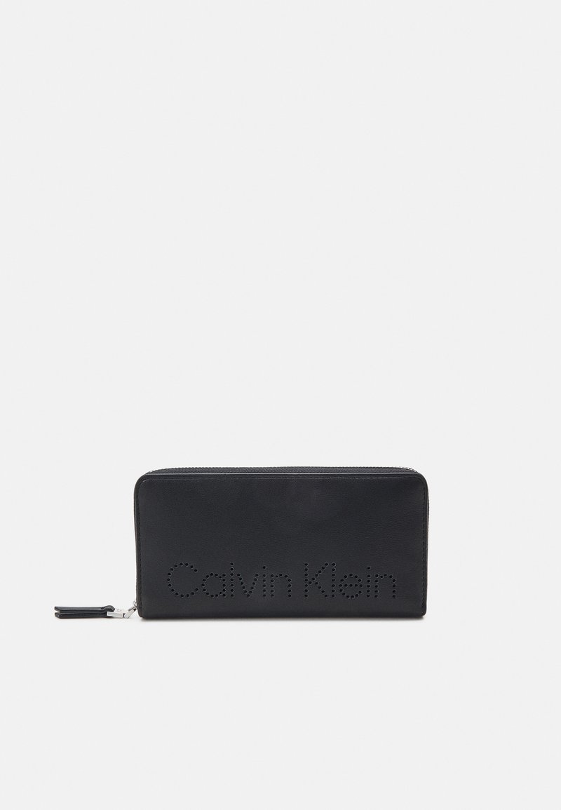 Crna kožna novčanica sa zatvaračem na patentni zatvarač. Sadrži perforirani logo "Calvin Klein" sprijeda i glatku teksturu. Tanak, pravokutni oblik.