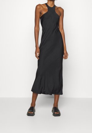 Vrouw draagt een mouwloze zwarte midi-jurk met een halternek en zwarte open-teen platformsandalen, staand tegen een effen witte achtergrond.