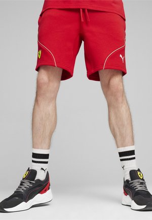 Czerwone spodenki sportowe z białymi detalami, z logo Ferrari. Połączone z czarnymi sneakersami i paskowanymi białymi skarpetkami.