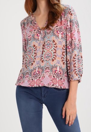 Femme portant un chemisier long à manches longues coloré à motifs avec un col en V, associé à un jean slim bleu foncé.