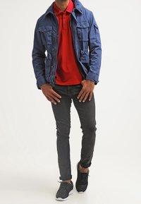 Homme portant une veste bleue par-dessus un polo rouge, un jean slim noir et des baskets noires sur fond blanc.