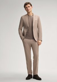 Lichtbruine pak met een getailleerde pasvorm, een enkelbreasted jasje, notch lapels en bijpassende broeken. Draag het over een bruine trui, gecombineerd met donkere laarzen.