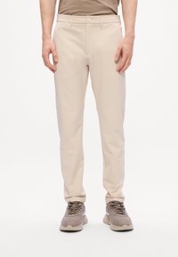 Man in beige rechte broek en taupe sneakers, staand met ontspannen handen langs zijn zij tegen een witte achtergrond.