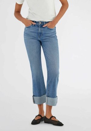 Persona che indossa jeans azzurri risvoltati, maglietta bianca a maniche corte e ballerine nere stile Mary Jane, in piedi con le mani nelle tasche.