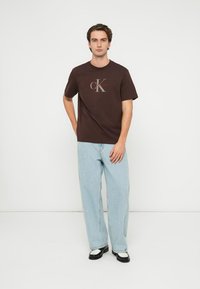 T-shirt de algodão castanha com logótipo CK, combinada com jeans azul claro de perna larga. Modelo usa sapatos pretos e brancos, a posar num fundo branco.