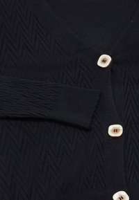 Černý cardigan s herringbone vzorem, s výstřihem do V, dlouhými rukávy a čtyřmi světlými knoflíky vpředu.