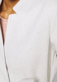 Jasnoszary, filcowy płaszcz z wysokim kołnierzem i eleganckimi liniami, charakteryzujący się minimalistycznym designem. Miękka tekstura bez widocznych zapięć.