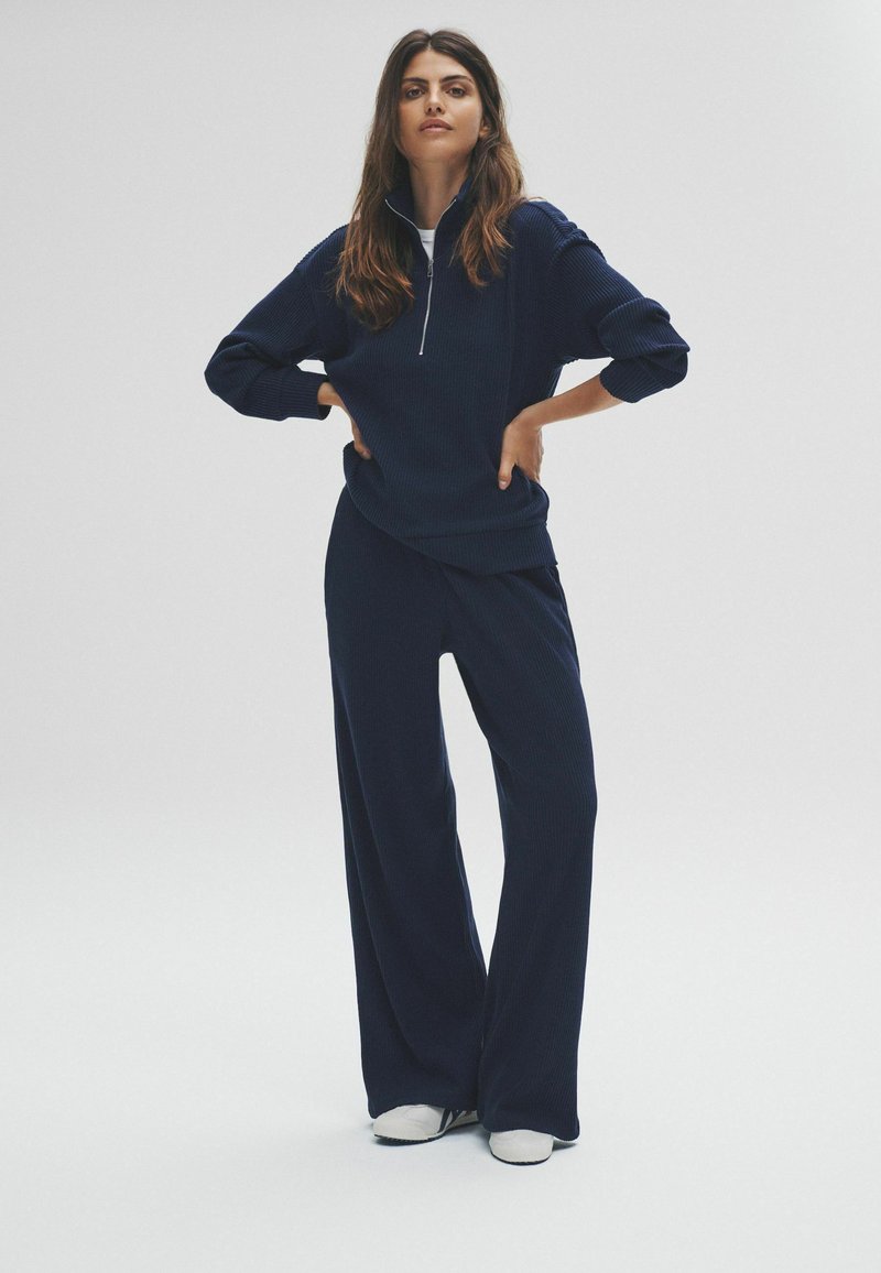 Femme debout avec les mains sur les hanches, portant un pull zippé côtelé bleu marine et un pantalon large, avec des baskets blanches sur un fond uni.