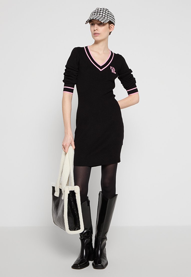 Frau in einem schwarzen Strickkleid mit rosa Akzenten, Hahnentrittmütze, schwarzen Strumpfhosen, hohen schwarzen Stiefeln und einer schwarz-weißen Tragetasche.
