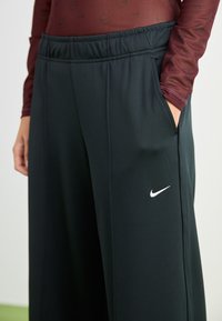 Sorte treningsbukser laget av glatt stoff med elastisk midjebånd, med sidelommer og en hvit Nike-logo på leggen.