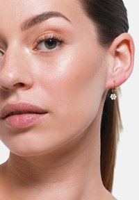 Orecchino a cerchio dorato con un design floreale e perline bianche, indossato su un tono di pelle chiara e liscia. Focus sui dettagli eleganti e sull'artigianato.