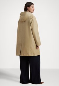 Μπεζ παλτό με κουκούλα και ίσια γραμμή, με πίσω σκίσιμο, μακριά μανίκια και απαλή υφή, συνδυασμένο με φαρδιά σκούρα παντελόνια.