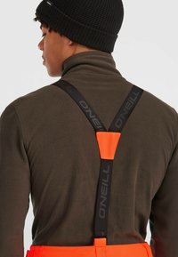 Orange hängslen med svart O'Neill-märke fästa vid en mörkbrun fleece-tröja, framhävd av en strukturerad svart stickad mössa.
