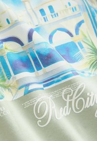 Cotton T-shirt featuring blue and green architectural illustration with palm leaves; "Red City" text in white at the bottom. Soft texture.