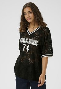 Mlada ženska z valovitimi rjavimi lasmi nosi črno čipkasto bluzo z napisom "COLLEGE 74" v belih črkah in temno modre kavbojke na enobarvni ozadju.