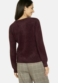 Maglione a maglia bordeaux con una consistenza morbida, scollatura rotonda e lunghe maniche a sbuffo. Abbinato a pantaloni con motivo a quadri.
