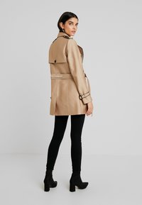 Beige, mid-length trench coat with a belted waist, buttoned tab collar, and contrasting black accents. Worn over black leggings and ankle boots.