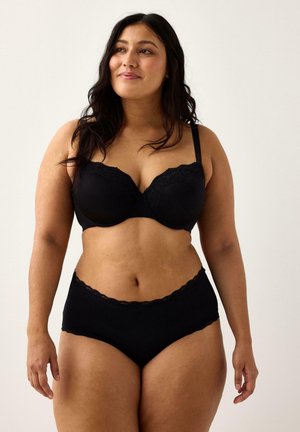 Donna con lunghi capelli scuri che indossa un reggiseno di pizzo nero e mutandine a vita alta coordinate, in piedi contro uno sfondo chiaro e semplice.