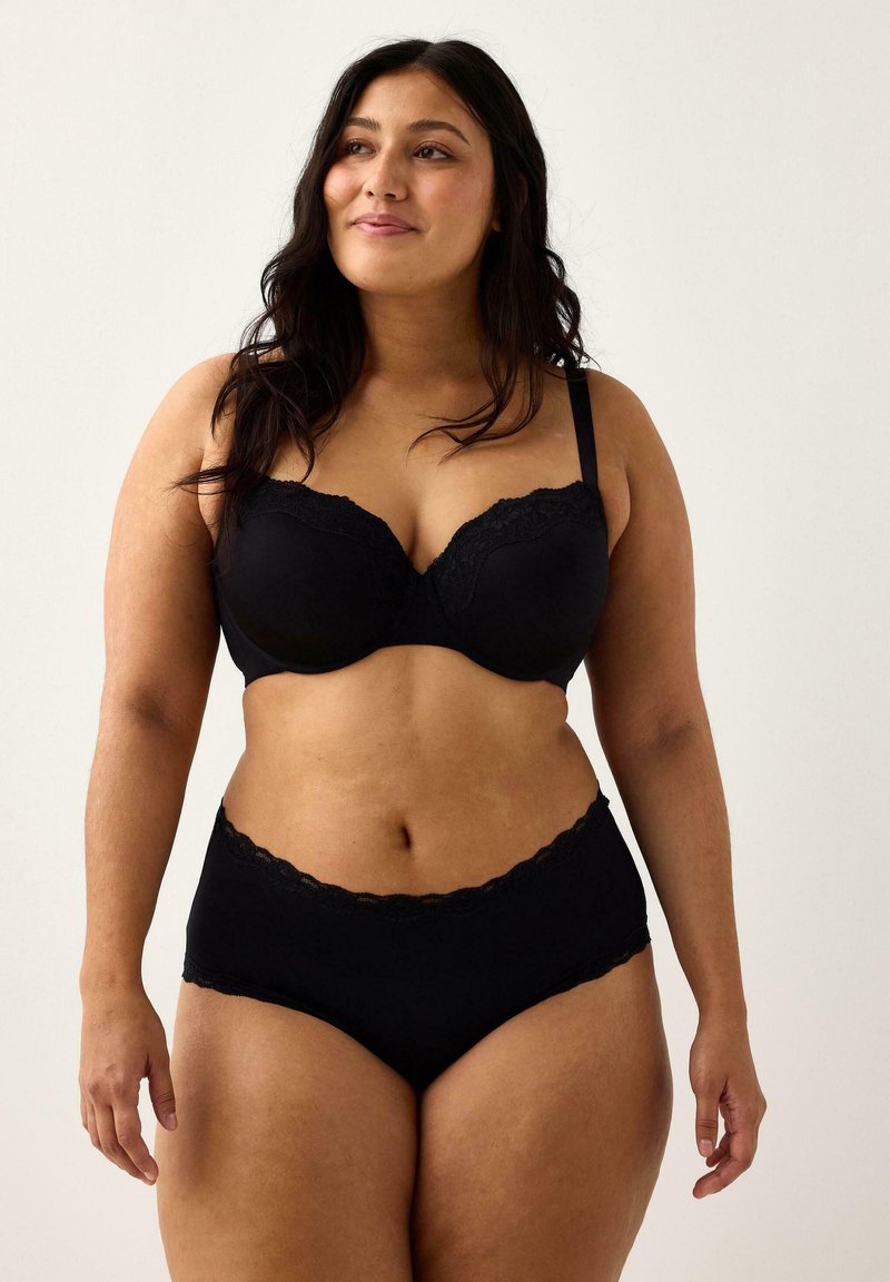 Kvinde med langt, mørkt hår iført en bh med sort blonder og matchende højtaljede trusser, stående foran en ensfarvet lys baggrund.