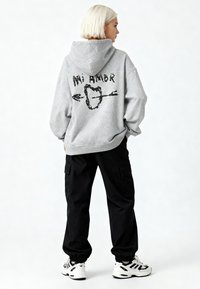 Pilkas gobtuvas su juoda grafika ant nugaros, kurioje užrašyta "Mi Amor." Dėvima su juodomis kargo kelnėmis ir baltomis sportinėmis bateliais.