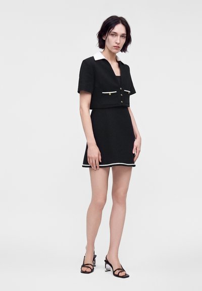Femme debout portant une veste courte noire à manches courtes avec un col blanc et une jupe noire assortie avec un liseré blanc et des sandales noires à talons.
