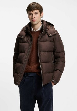 Joven con una chaqueta acolchada de color marrón oscuro con bolsillos con cremallera, un suéter de punto grueso color naranja quemado y pantalones de pana azul oscuro.