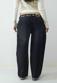 Kobieta w szerokich czarnych dżinsach z tylnymi kieszeniami, z dżinsowym paskiem z ćwiekami, kremowym swetrem prążkowanym i srebrnymi butami, stojąca tyłem.