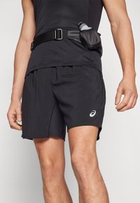 Schwarze sportliche Shorts aus einem leichten Stoff. Mit einem seitlichen Schlitz am Saum und einem Logo auf der linken unteren Seite. Begleitet von einem Gürtel mit einer Wasserflasche.